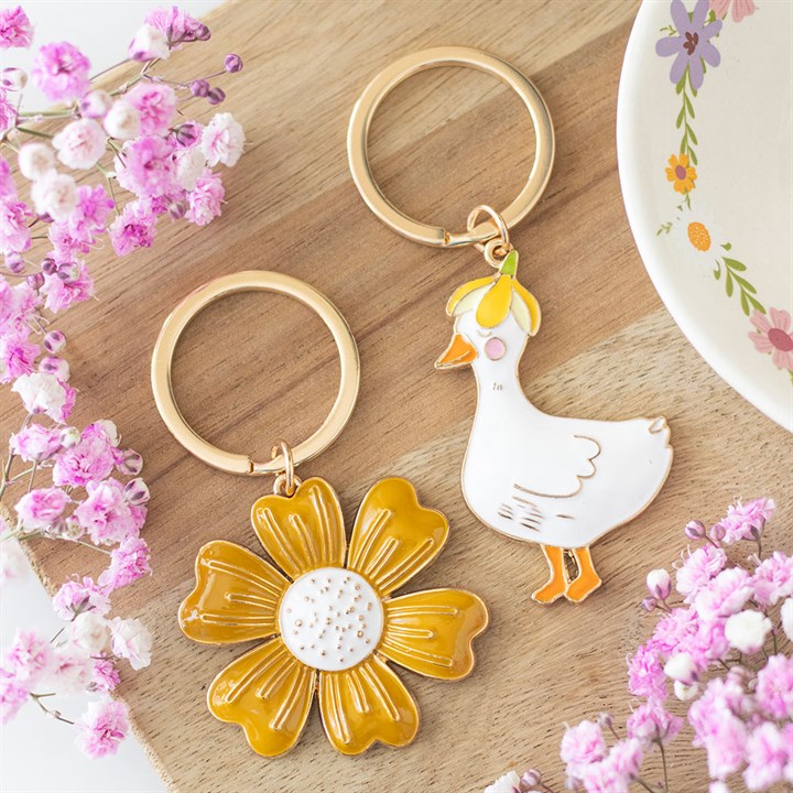 Two keychains, one with a yellow flower and the other with a white duck design, on a wooden surface with pink flowers.