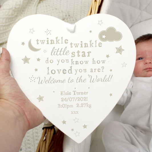 A large wooden heart-shaped decoration with the text 'Twinkle twinkle little star do you know how loved you are' and other stars and a cloud, held by a hand with a baby in the background.