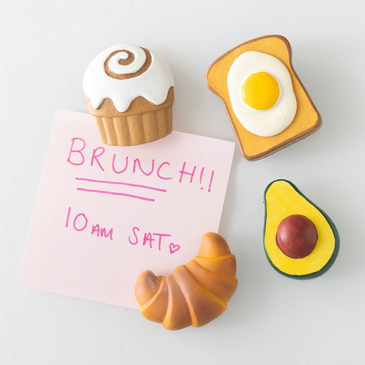 Fridge magnets shaped like a croissant, egg, cupcake, and avocado holding up a post-it note with 'BRUNCH!! 10 AM SAT' text.