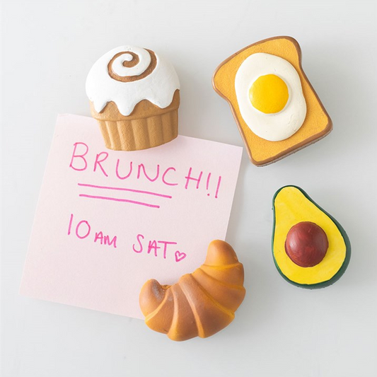 Fridge magnets shaped like a croissant, egg, cupcake, and avocado holding up a post-it note with 'BRUNCH!! 10 AM SAT' text.