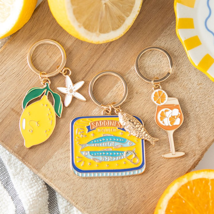 Three keychains a lemon, a sardine tin and cocktail glass designs on a wooden surface.