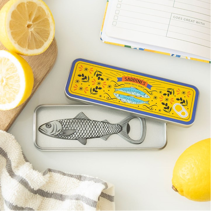 A silver fish-shaped bottle opener resting in its yellow and blue illustrated tin, styled on a white surface with lemon slices, a striped cloth, and an open notebook.