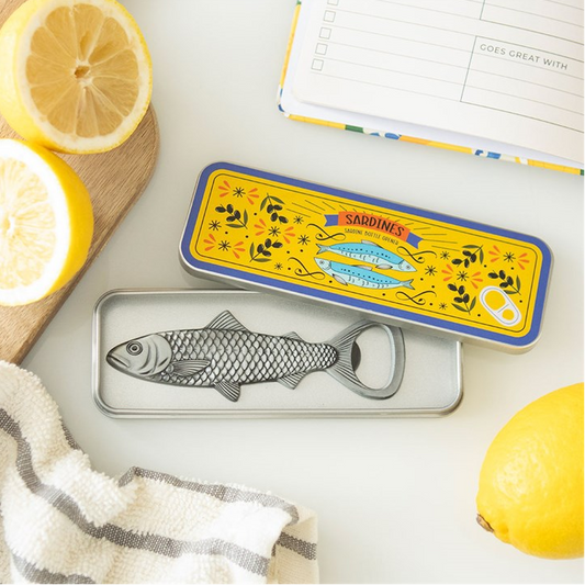 A silver fish-shaped bottle opener resting in its yellow and blue illustrated tin, styled on a white surface with lemon slices, a striped cloth, and an open notebook.