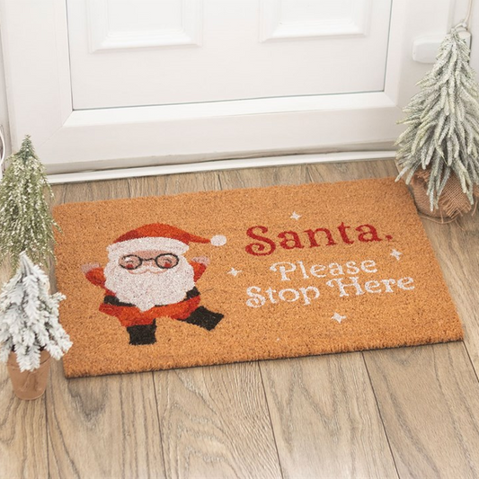 Doormat with Santa Claus design and text 'Santa, Please Stop Here' on a wooden floor.