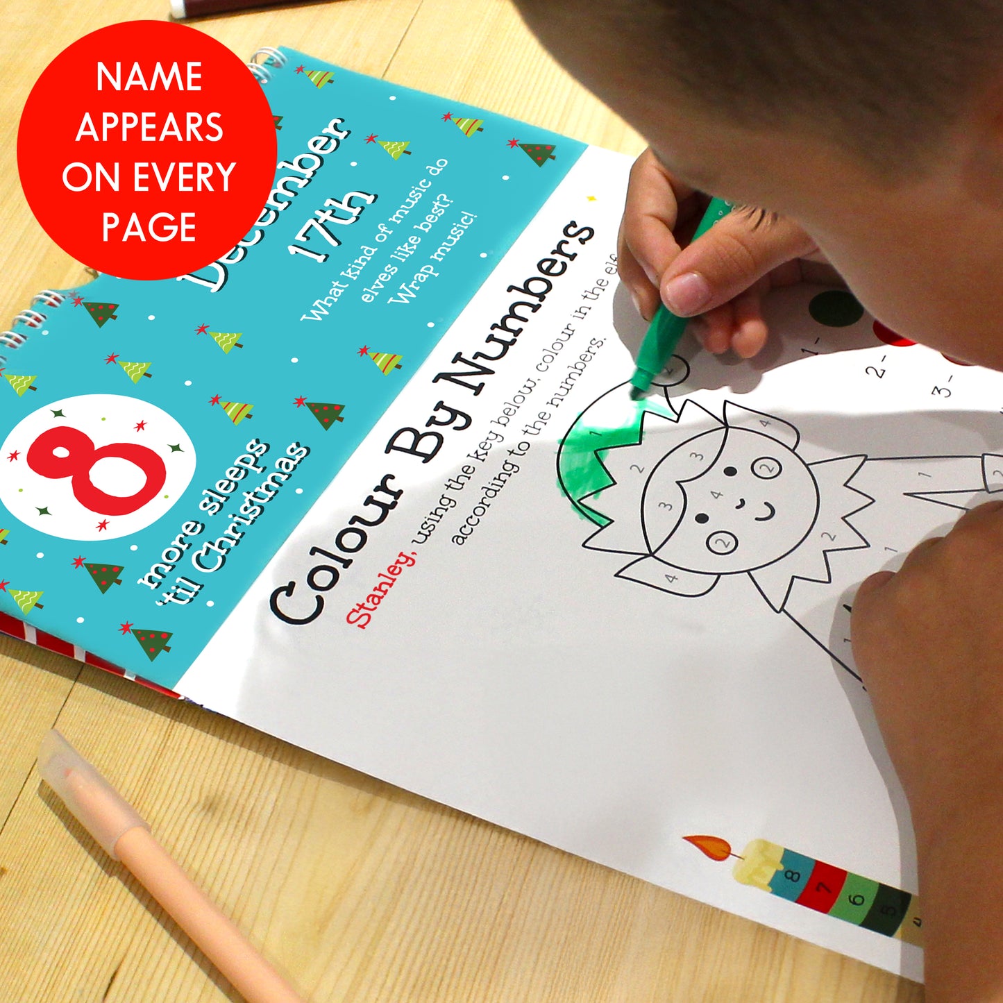 Child coloring a Christmas-themed book with a green crayon on a wooden surface.