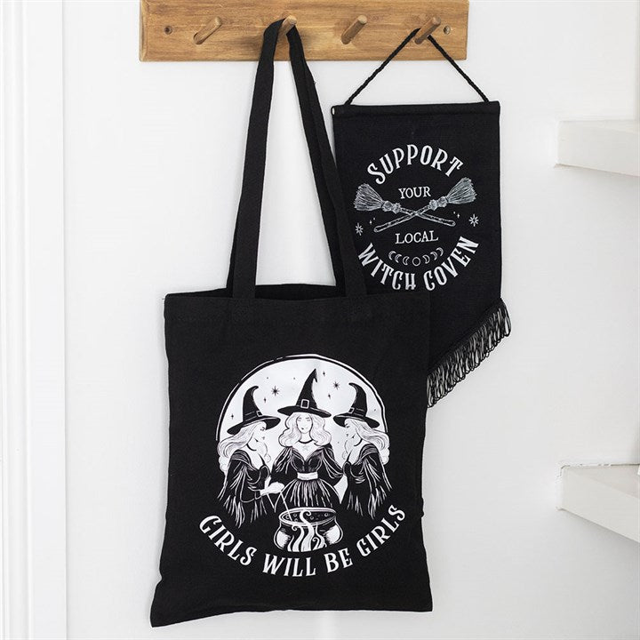 Black tote bag with witch-themed design and text, hanging on a wooden hook.