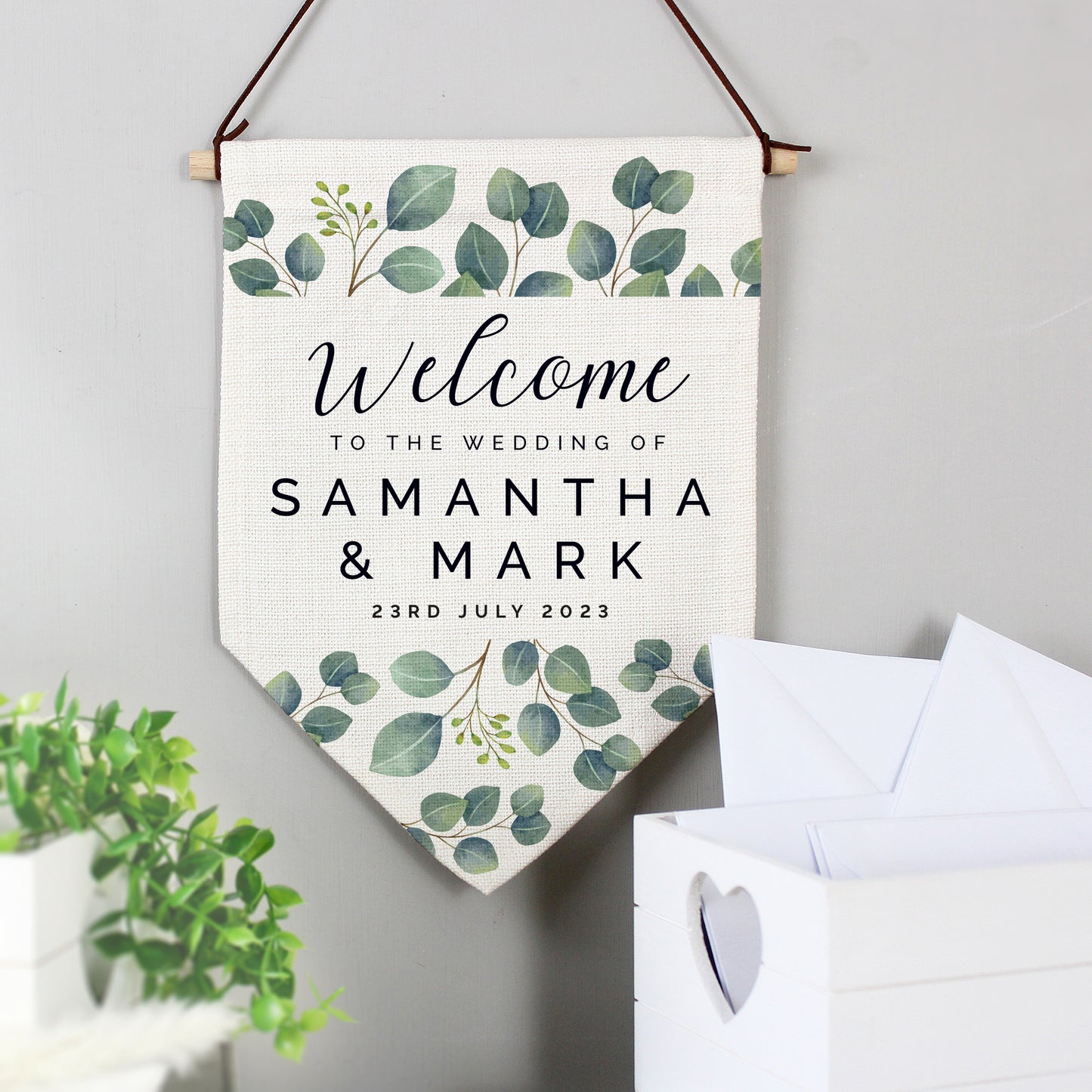 A personalised botanical wedding hanging banner with text 'Welcome to the Wedding of Samuel & Mark' and a date '23rd July 2023' on it, displayed above a card box.