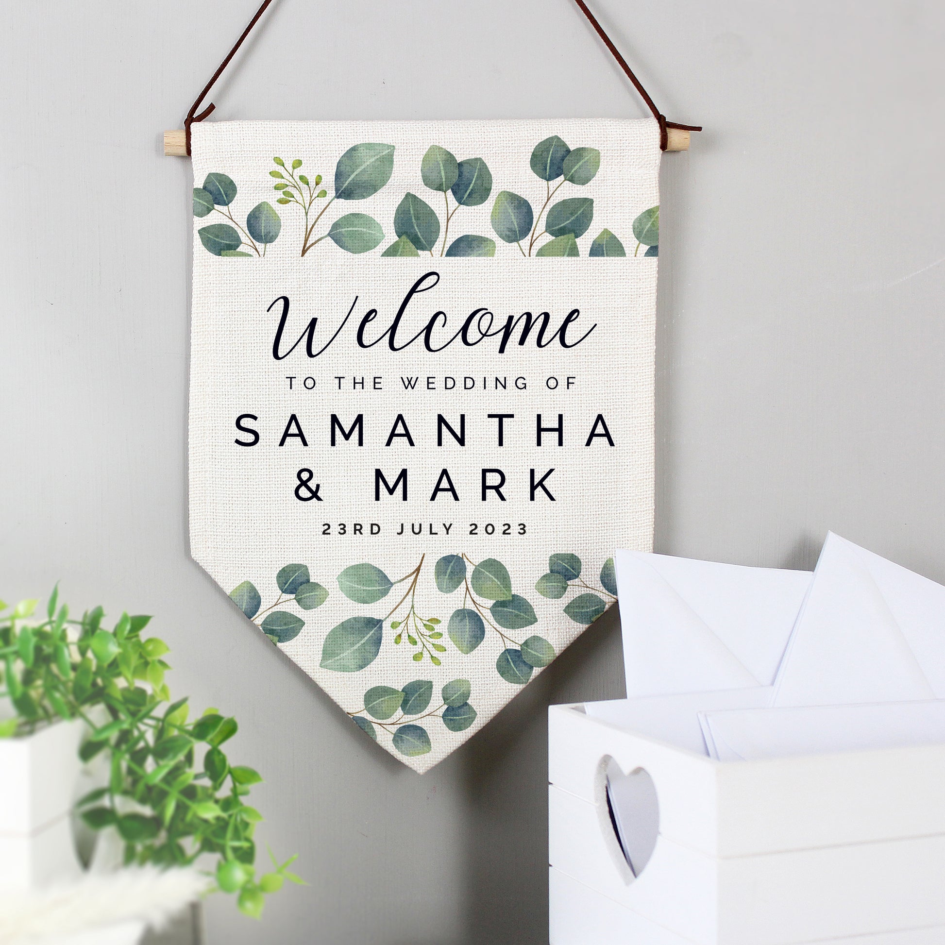 A personalised botanical wedding hanging banner with text 'Welcome to the Wedding of Samuel & Mark' and a date '23rd July 2023' on it, displayed above a card box.