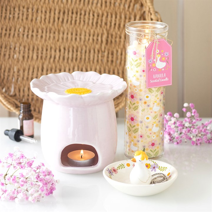 Pink daisy flower wax warmer with a lit candle, jar of vanilla-scented candle, and small duck-shaped container on a table with flowers.