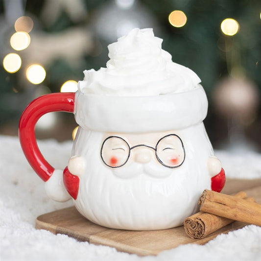 Ceramic mug shaped like a cartoon Santa character with a red handle, surrounded by cinnamon sticks on a snowy background.