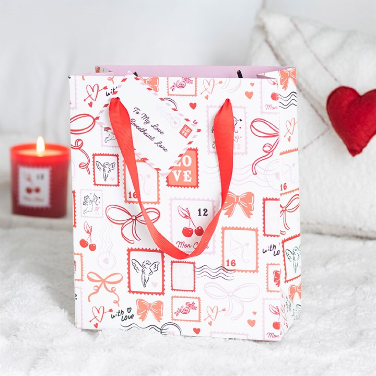 Gift bag with red handles and decorative patterns on a white surface with a candle and heart-shaped object in the background.