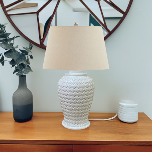 A woven ceramic table lamp with a geometric pattern in a beige color, featuring a linen shade on a wooden table.